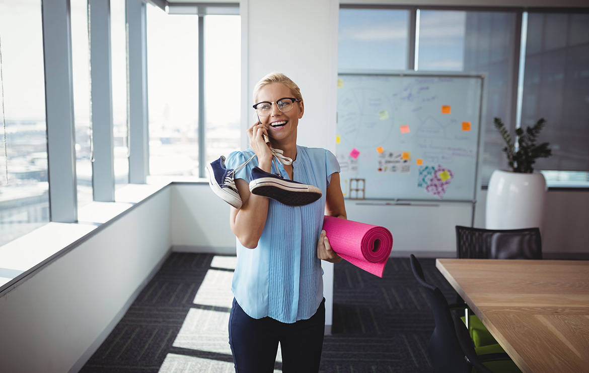 Smiling woman holding gym mat and shoes while on the phone