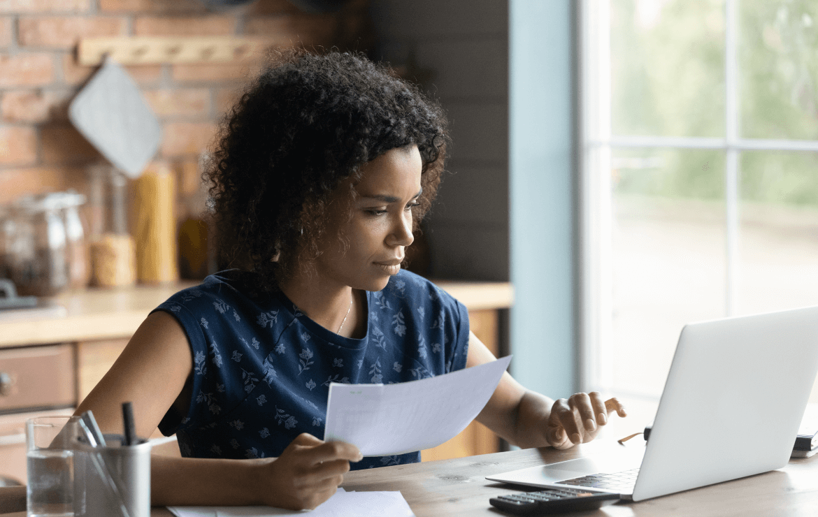 Woman holding piece of paper and scrolling on laptop