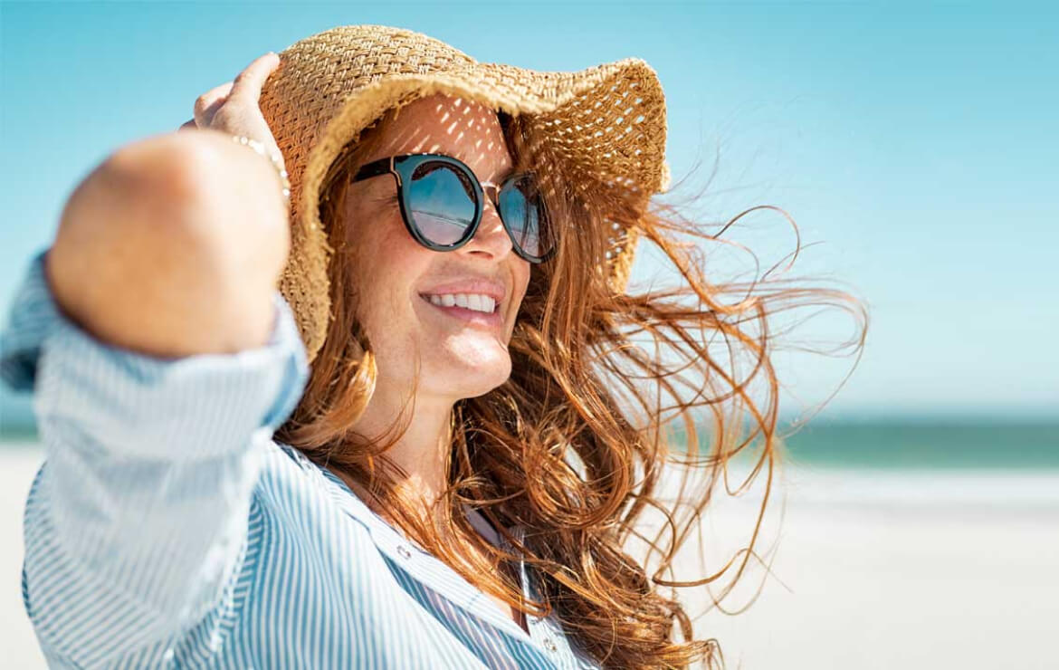 Woman smiling while sitting on the beach holding down hat
