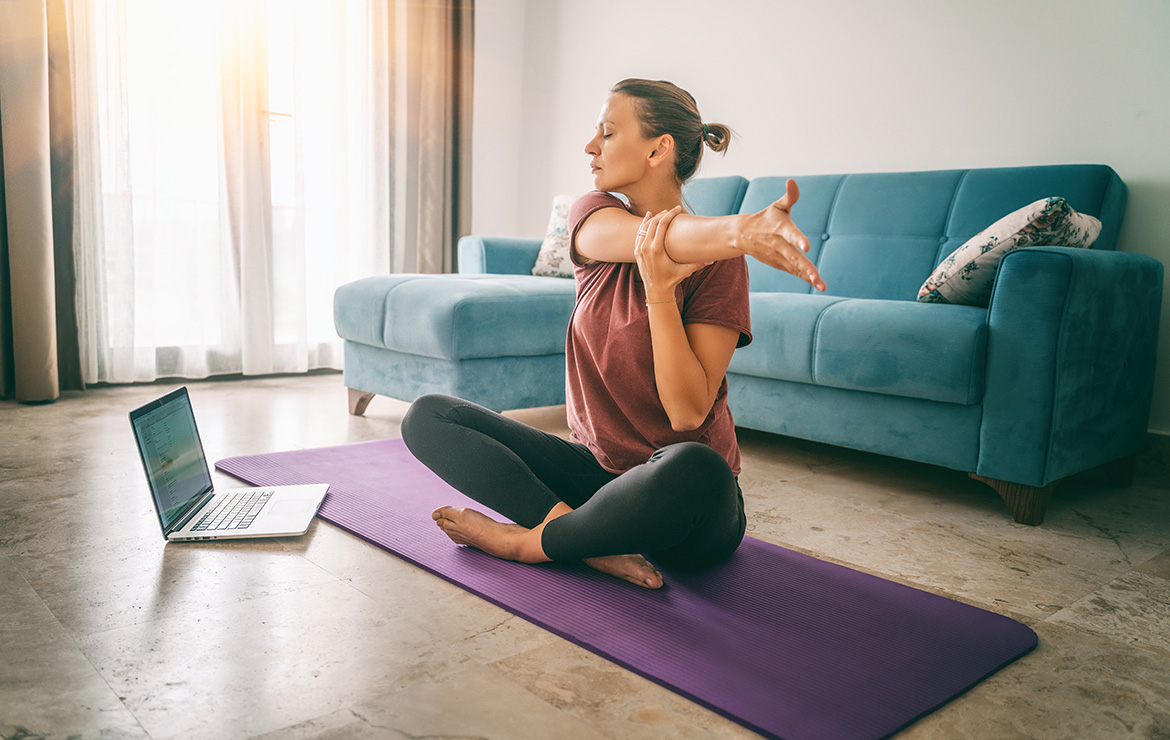 Woman sitting on yoga mat stretching and watching laptop
